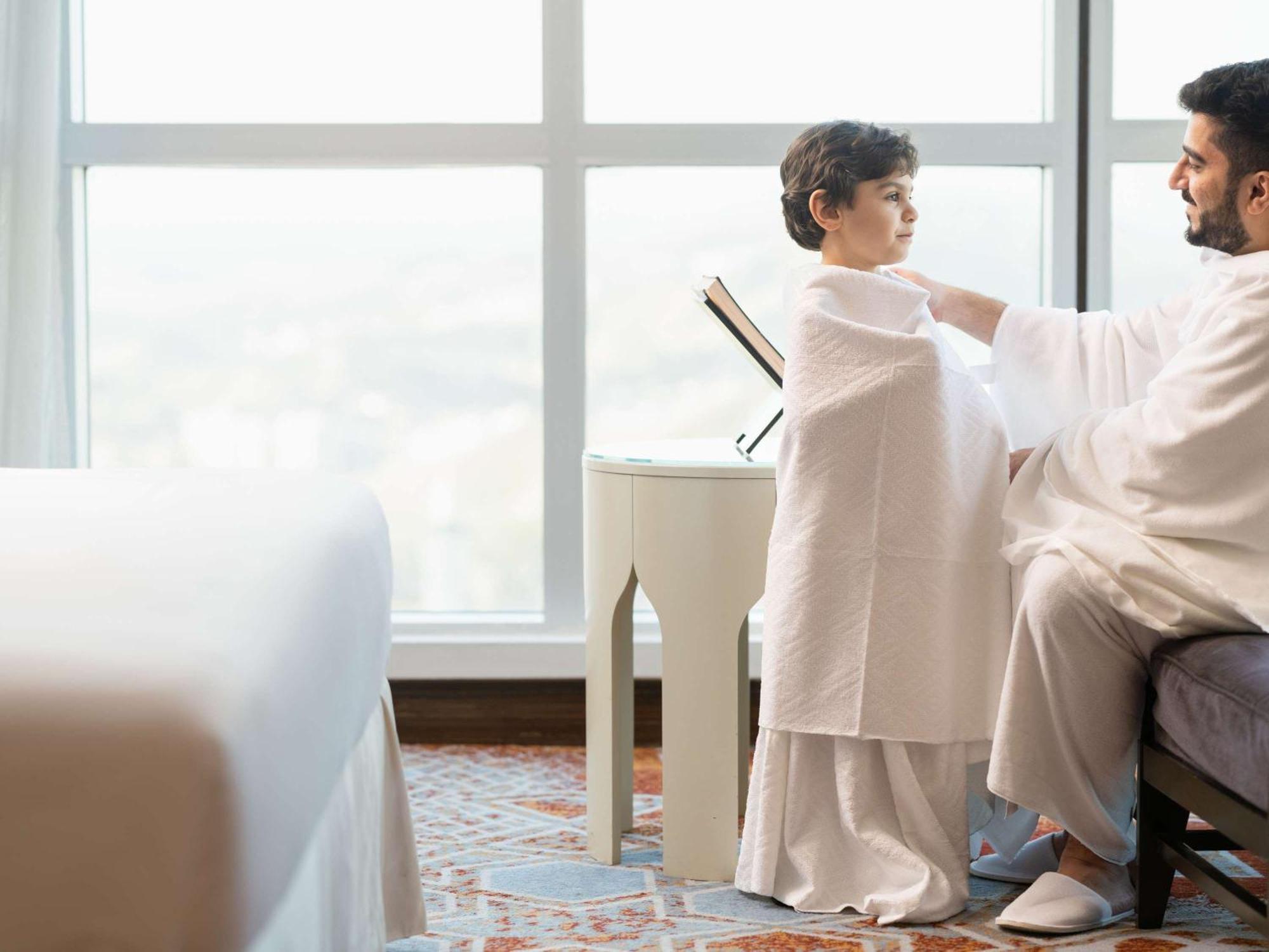 Swissotel Makkah La Meca Exterior foto The photo shows a man and a child in a bright, contemporary hotel room. The man, dressed in traditional attire, is gently assisting or guiding the child, who is also wrapped in a similar garment. They are seated near a small table by a large window, 