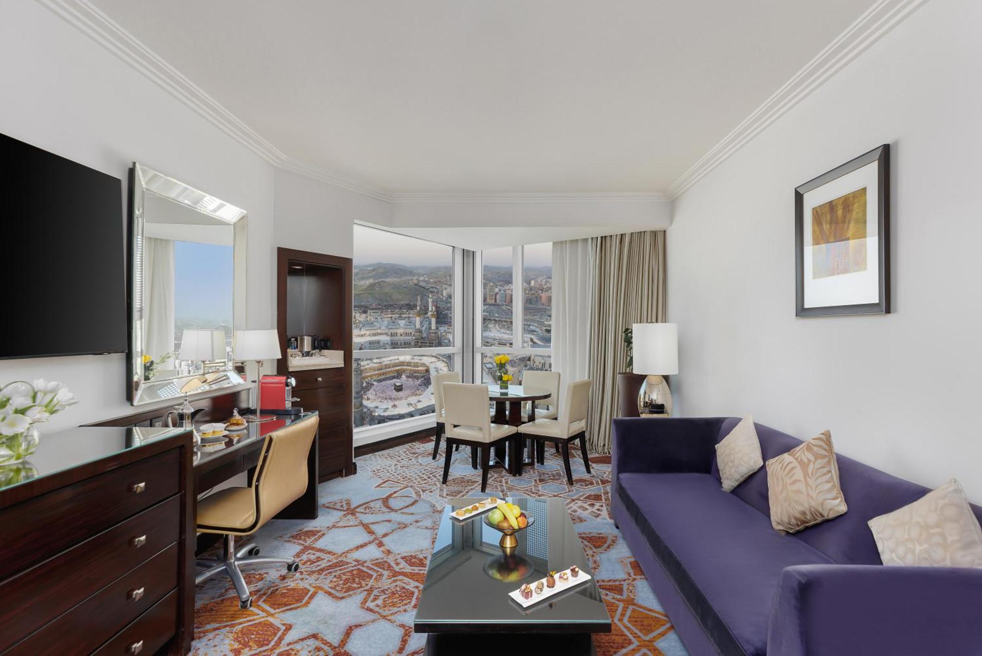 Swissotel Makkah La Meca Exterior foto The photo shows a hotel room with a modern and stylish design. There is a comfortable purple sofa in the foreground, facing a small coffee table with some decorative items on it. Near the sofa, there is a work desk with a chair and a flat-screen TV m