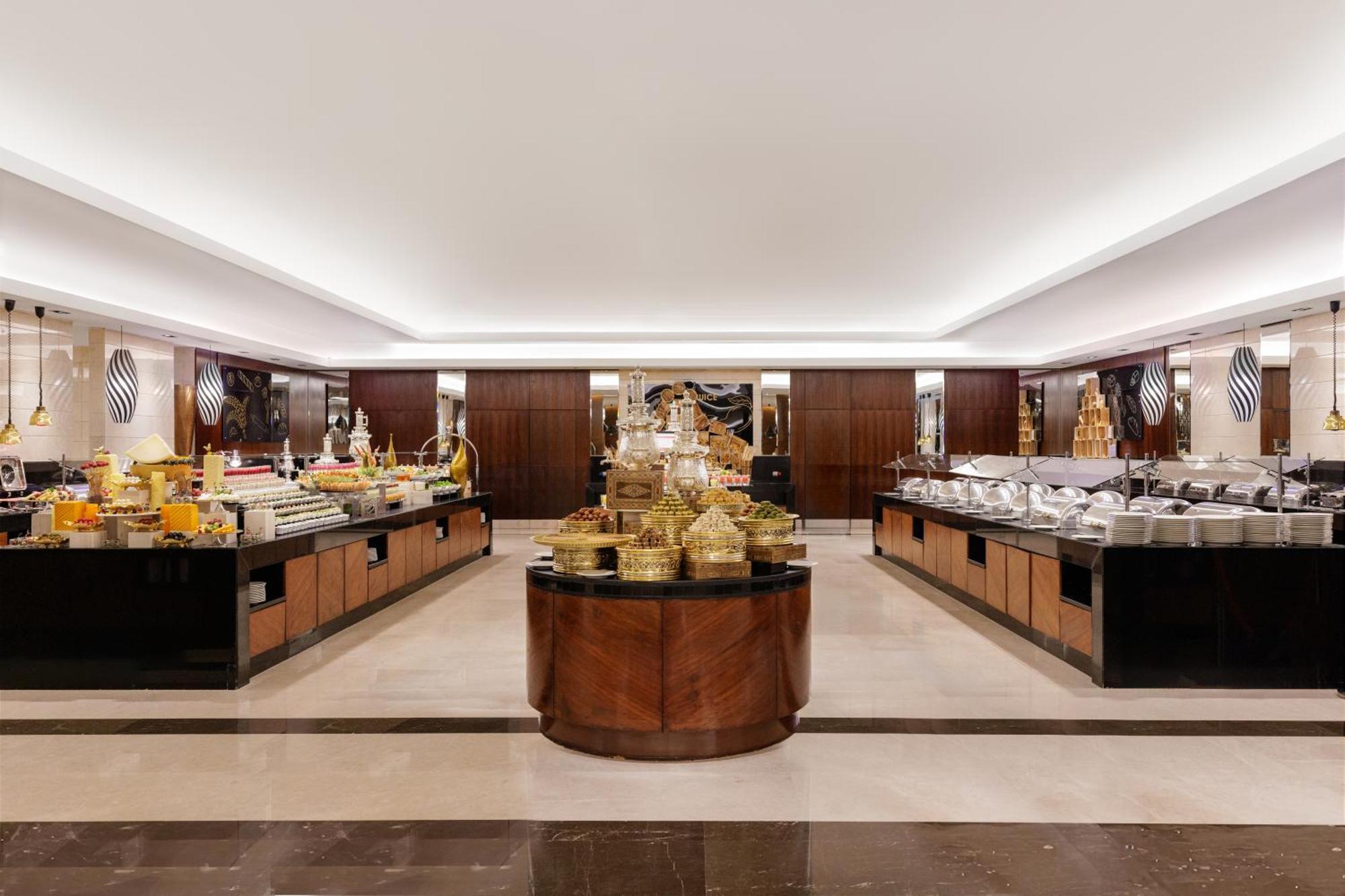 Swissotel Makkah La Meca Exterior foto The photo shows a spacious buffet dining area. The setting features elegant decor with polished surfaces and warm lighting. In the center, there is a large circular display of intricate food containers, likely showcasing an assortment of dishes. Surr