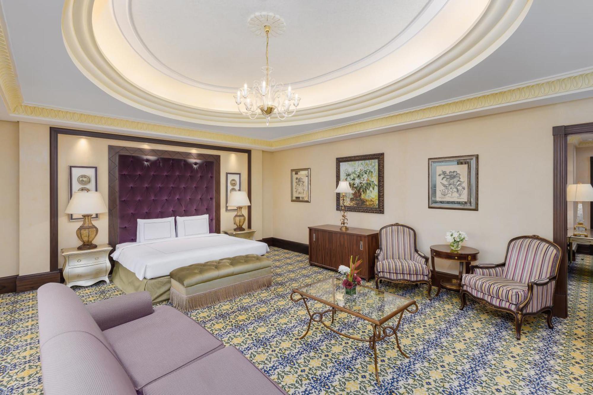 Swissotel Makkah La Meca Exterior foto The photo shows an elegantly designed hotel room. It features a large bed with a tufted purple headboard, complemented by two bedside lamps. There is a stylish sofa along one side, offering a comfortable seating area. Across from the bed, there is a 