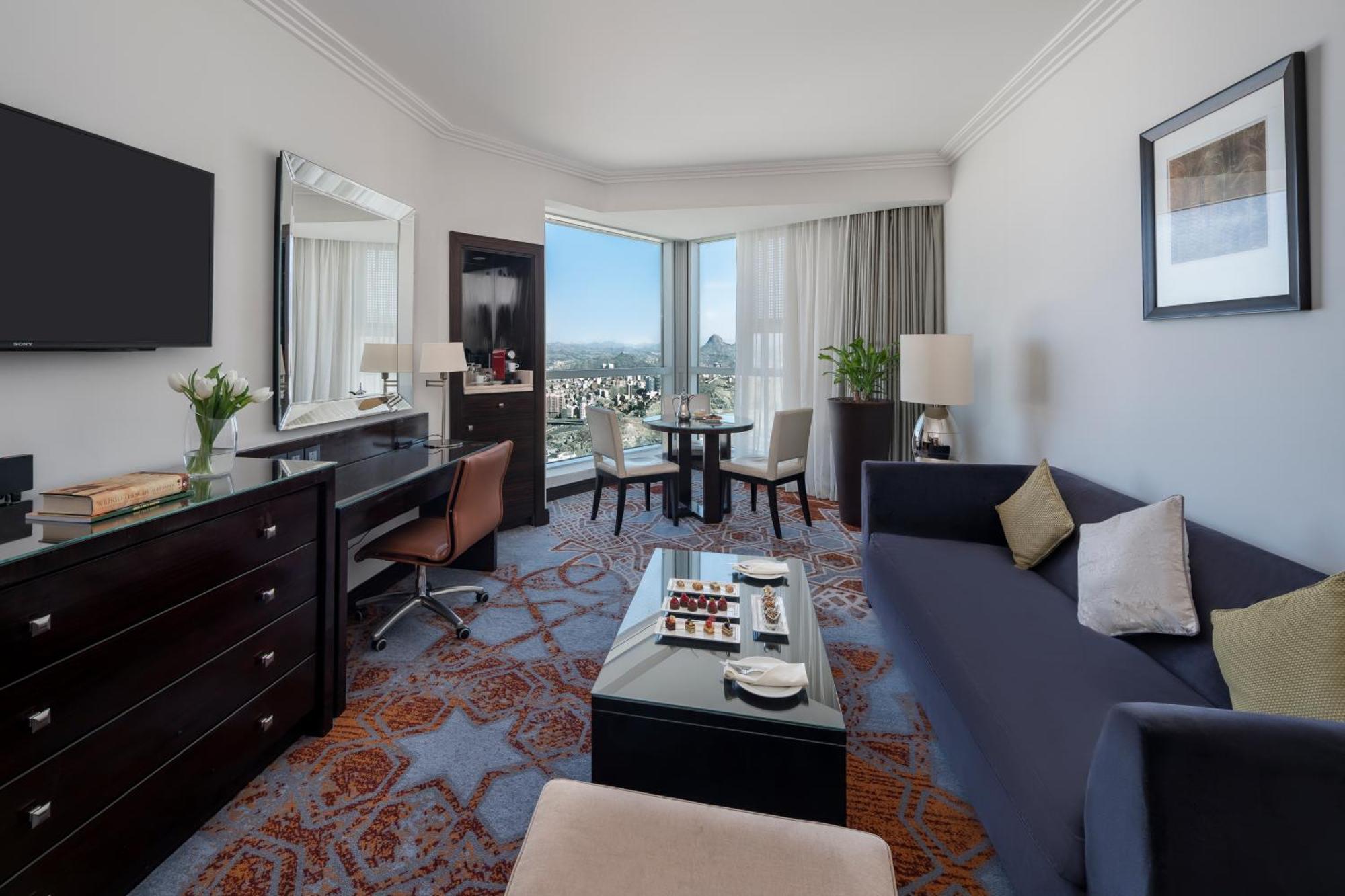 Swissotel Makkah La Meca Exterior foto The photo shows a well-furnished hotel room or suite. It features a comfortable dark blue sofa with decorative pillows, a glass coffee table set in front of it that has some snacks or refreshments. There's a stylish desk with a chair, likely for work