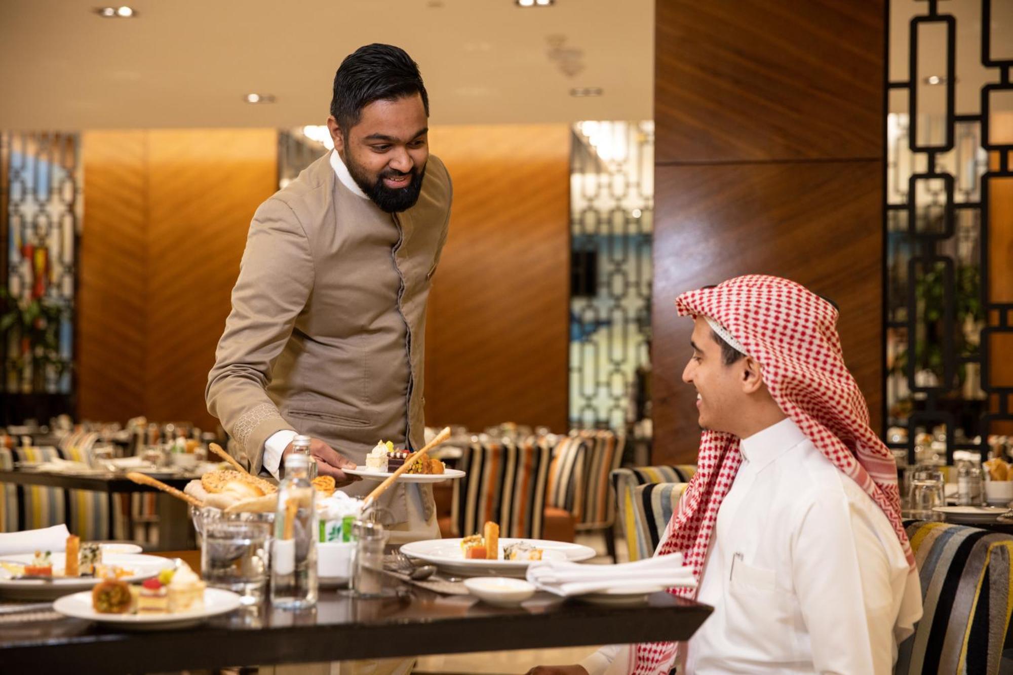 Swissotel Makkah La Meca Exterior foto The photo shows a scene in a restaurant where a waiter is serving a meal to a diner. The waiter, dressed in a formal outfit, appears to be presenting a dish with a smile. The diner, wearing traditional attire, looks engaged and attentive. The dining 