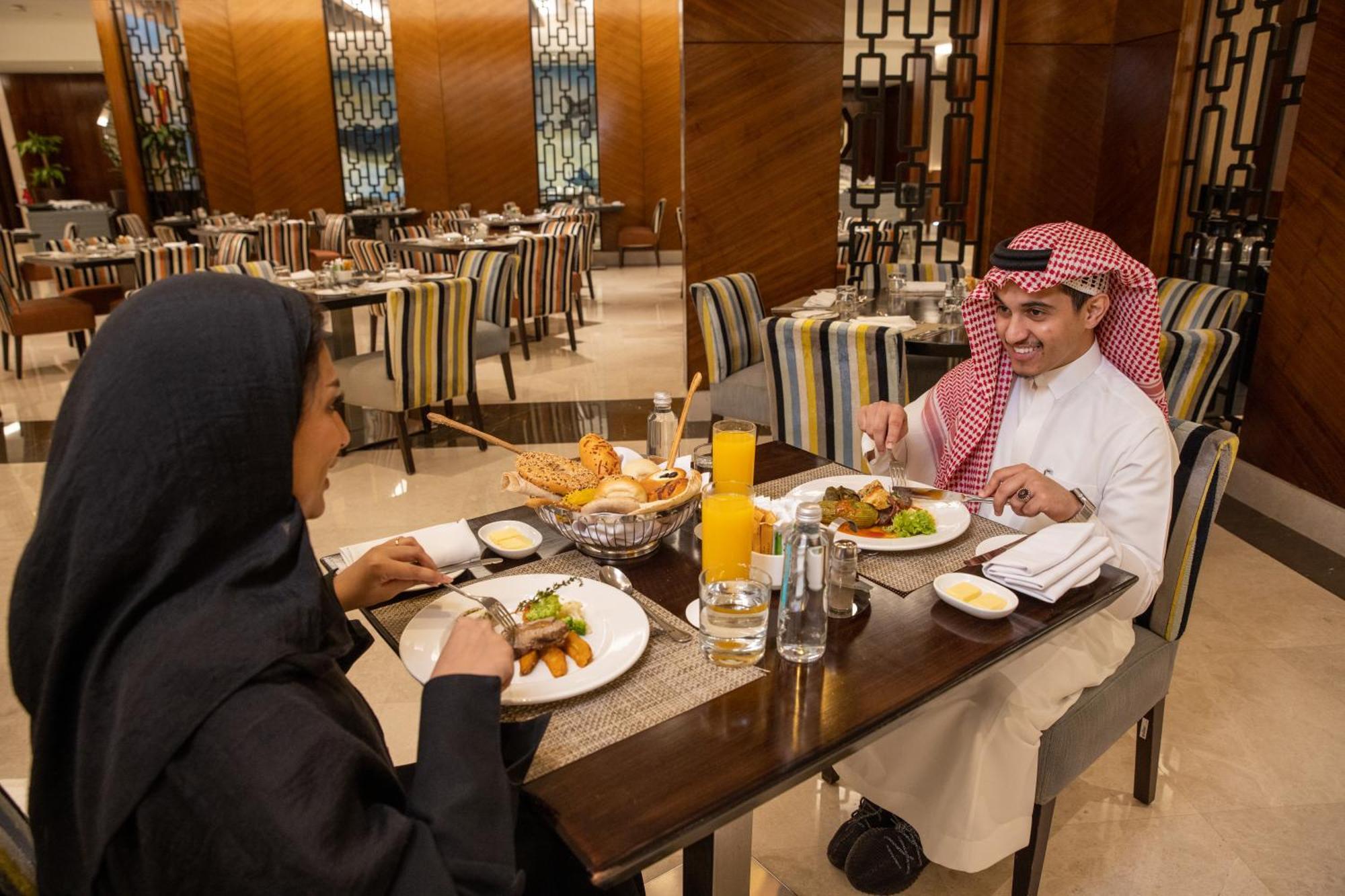 Swissotel Makkah La Meca Exterior foto The photo depicts a dining scene in a restaurant. It features two individuals seated at a table engaging in a meal together. One person is dressed in traditional attire, while the other is wearing a black hijab. The table is set with plates of food, 