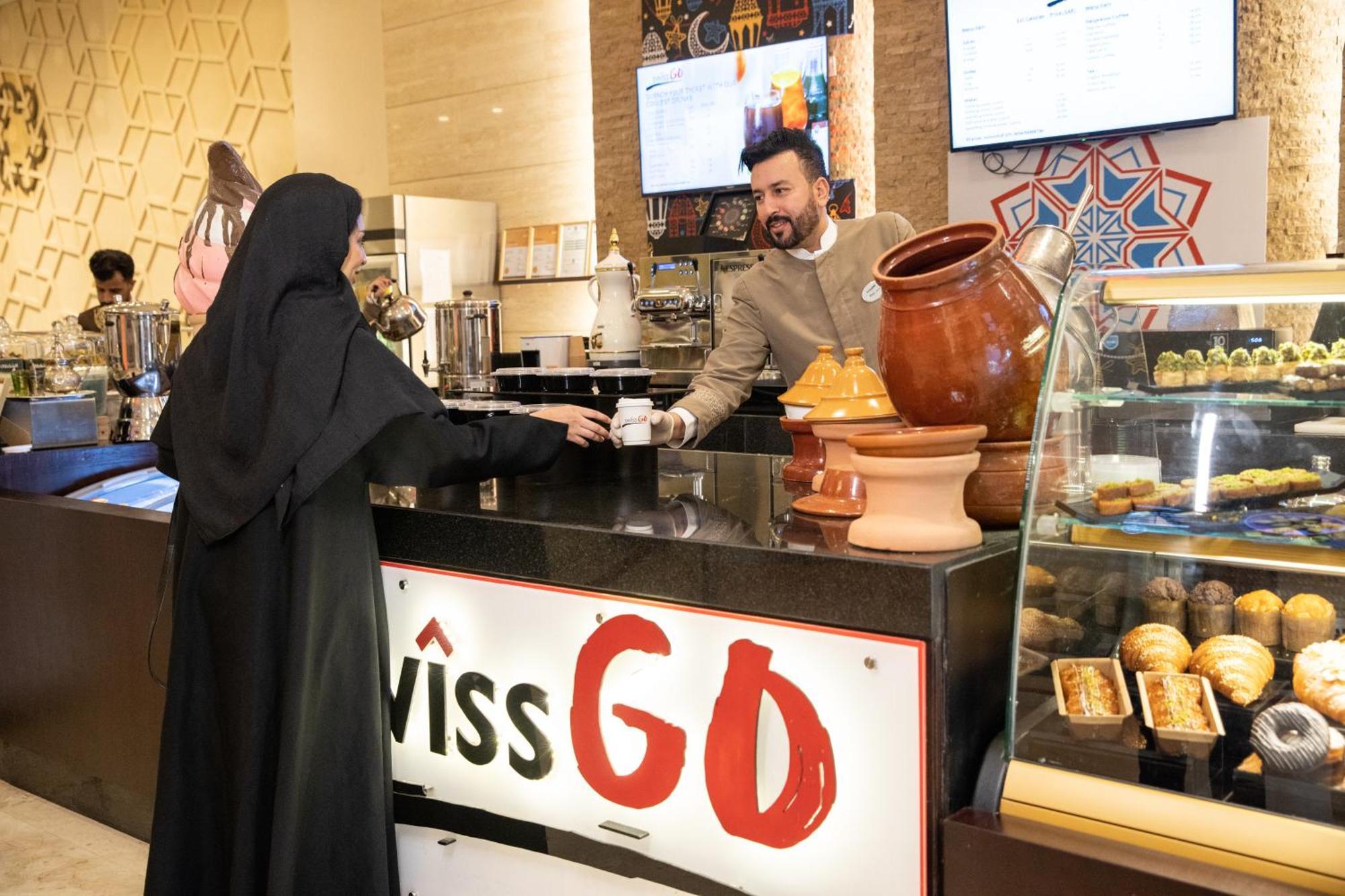 Swissotel Makkah La Meca Exterior foto The photo shows a scene inside a cafe or restaurant. A man in a beige outfit is serving a woman who is wearing a black traditional dress, possibly an abaya. The setting appears modern and includes decorative elements, such as patterned wall decoratio