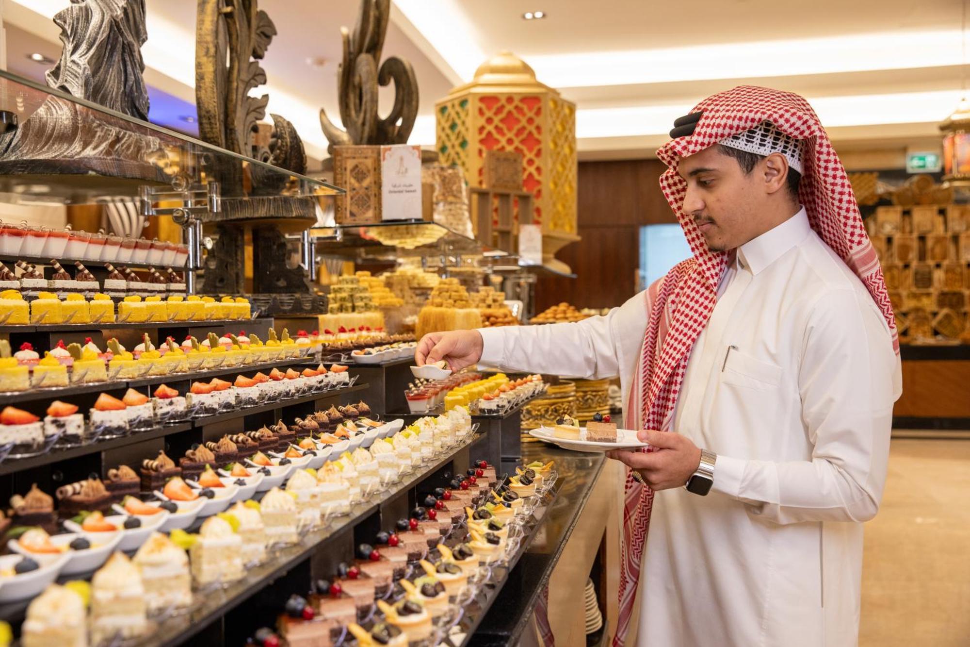 Swissotel Makkah La Meca Exterior foto The photo shows a man dressed in traditional attire, wearing a white thobe and a red-and-white checked ghutrah (headscarf). He is standing in front of an elaborate display of desserts in a luxurious setting. The dessert display features a variety of 