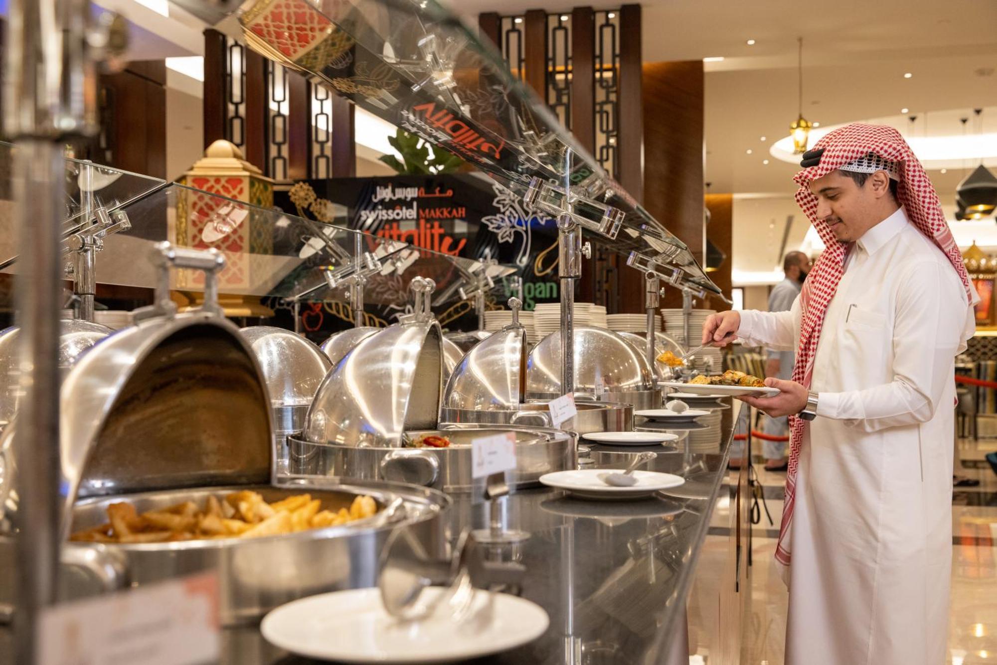 Swissotel Makkah La Meca Exterior foto The photo shows a buffet-style dining setup. A man dressed in traditional attire is standing in front of a row of shiny silver food warmers. He appears to be selecting food from the buffet. The background has decorative elements, and you can see vari