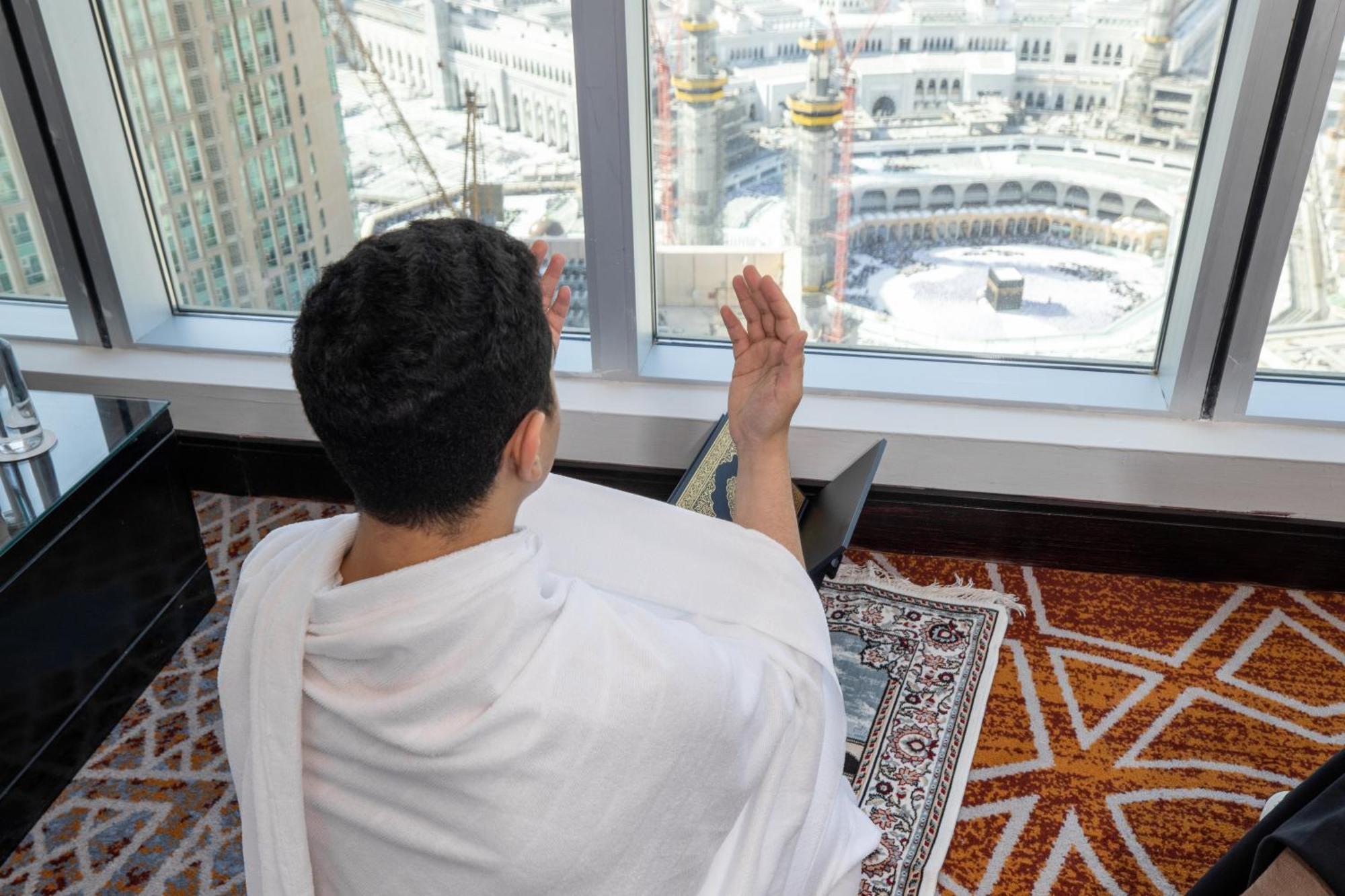 Swissotel Makkah La Meca Exterior foto The photo shows a person in traditional clothing, likely a Ihram, sitting in a room with a large window that overlooks a holy site, presumably the Kaaba in Mecca. The individual is engaged in prayer, raising their hands in a gesture commonly associat
