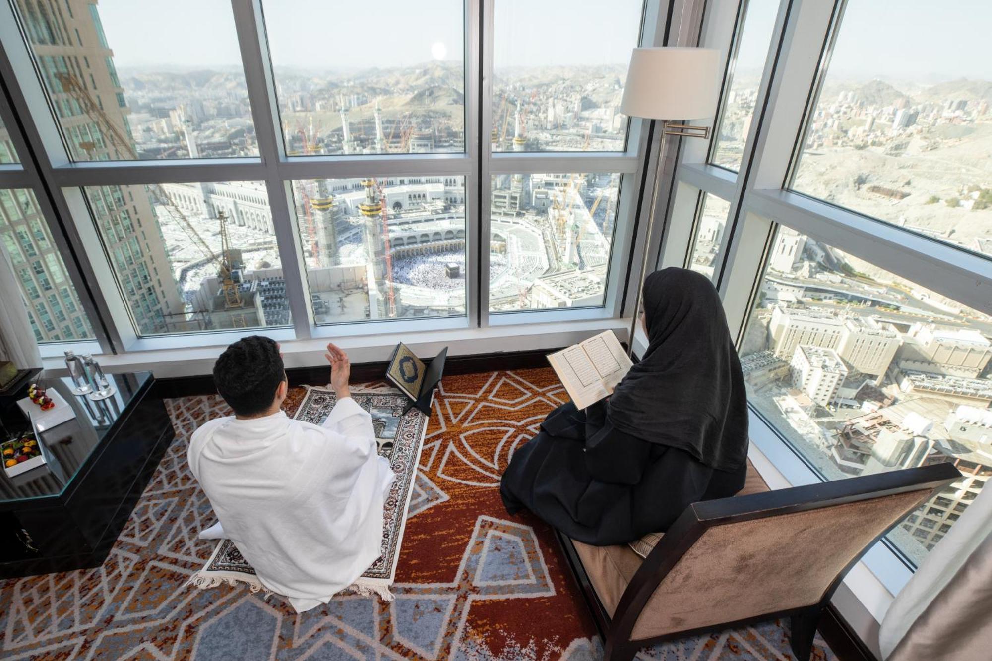 Swissotel Makkah La Meca Exterior foto The photo shows a man and a woman engaged in prayer and reading, respectively, inside a room with large windows offering a view of a cityscape. The man is dressed in traditional attire, facing a direction, possibly in a state of supplication. The wom