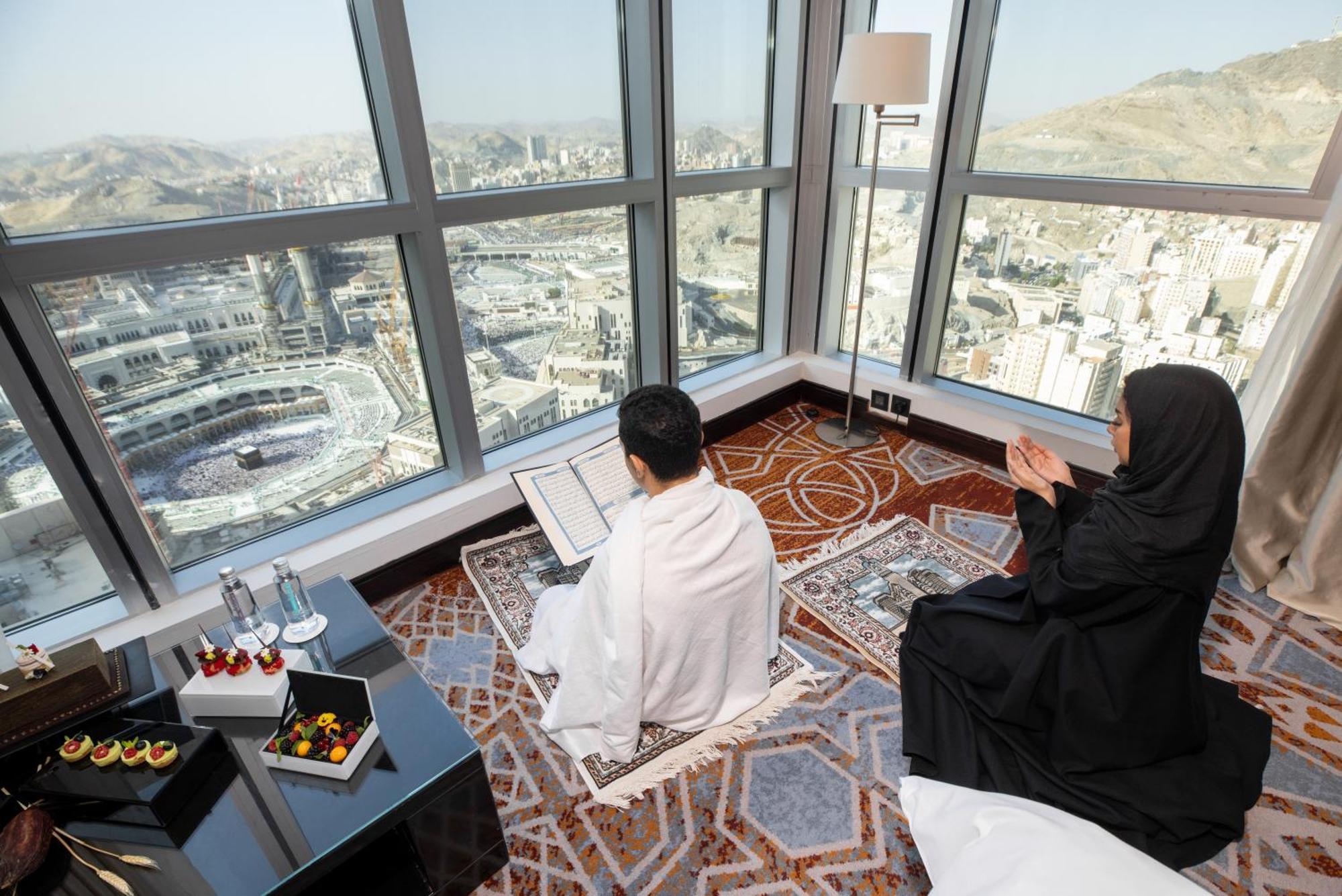 Swissotel Makkah La Meca Exterior foto The photo shows a serene interior scene with a view of a city, likely Mecca, through large windows. In the foreground, a person dressed in traditional white garments (Ihram) is seated on the floor, possibly reading or reciting from a book. Beside the