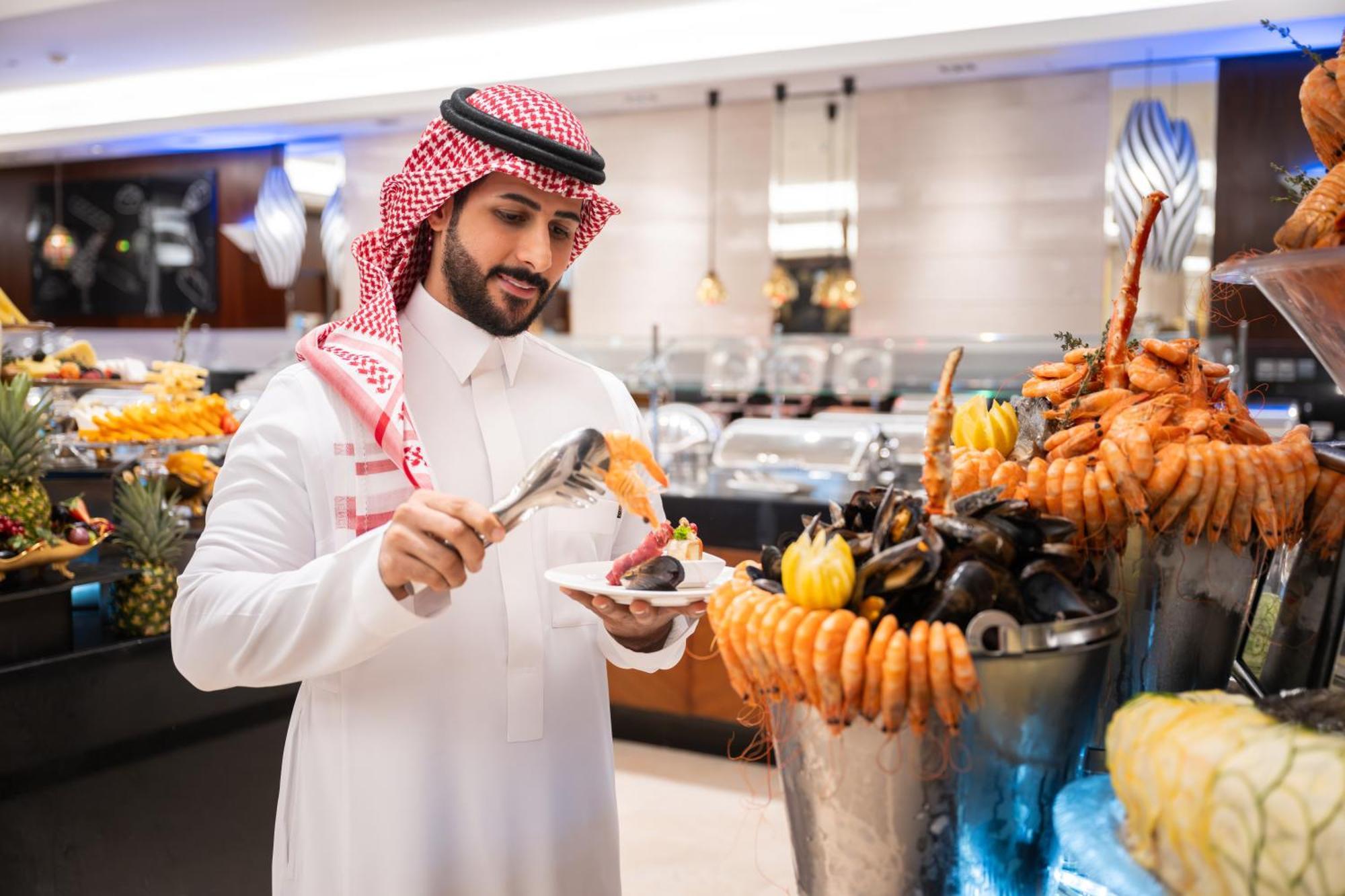 Swissotel Makkah La Meca Exterior foto The photo depicts a man dressed in traditional attire, appearing to be in a restaurant or buffet setting that features a lavish display of seafood. He is using tongs to serve food onto his plate from a large assortment of seafood, which includes item