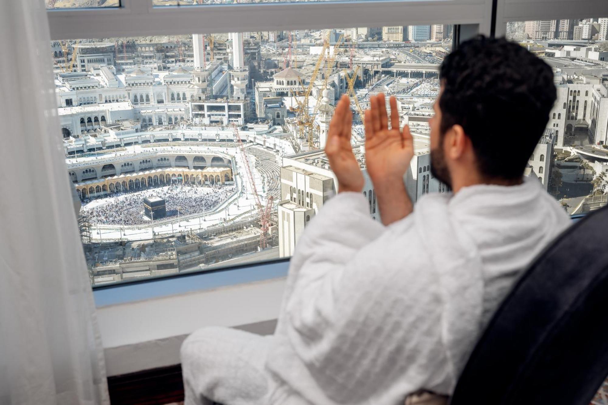 Swissotel Makkah La Meca Exterior foto The photo shows a person wearing a traditional white garment, seated by a large window with a view of a significant religious site. They appear to be in an attitude of prayer or supplication, looking out towards a vast area filled with many people. T