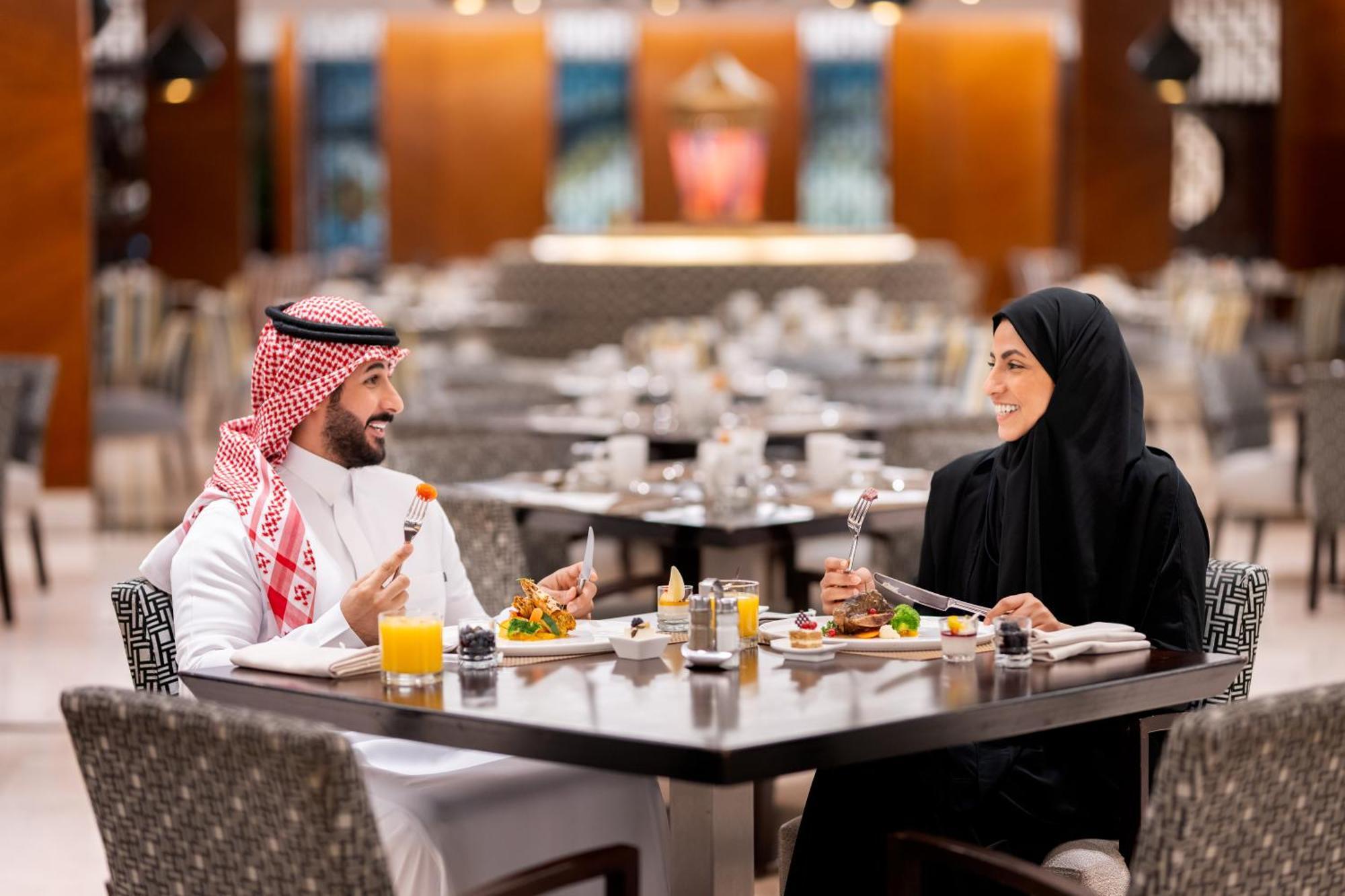 Swissotel Makkah La Meca Exterior foto The photo depicts a couple dining together at a restaurant. The man is dressed in traditional Saudi attire, which includes a white thobe and a red-and-white checked ghutrah (headscarf). The woman is wearing a black abaya and hijab. They are seated at