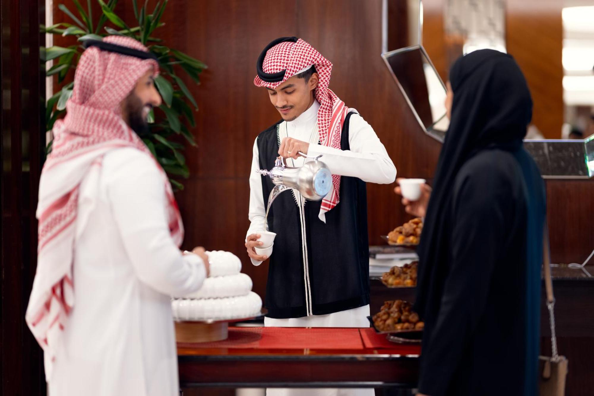 Swissotel Makkah La Meca Exterior foto The photo depicts a scene in a setting that resembles a formal gathering or event. In the foreground, a man in traditional attire, wearing a white thobe with a red and white checked headscarf, is pouring a beverage from a silver coffee pot into a sma