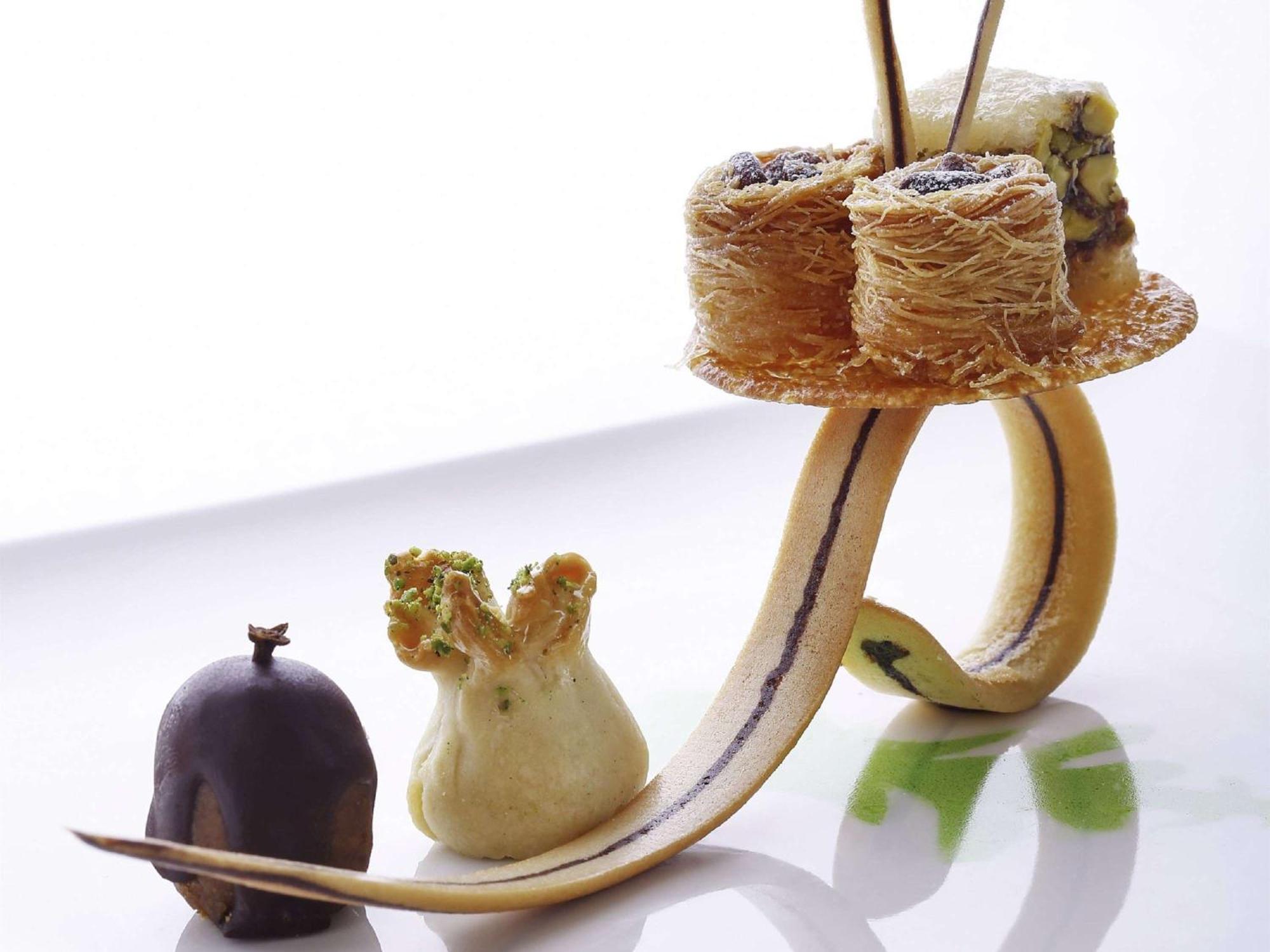 Swissotel Makkah La Meca Exterior foto The photo shows an artistic dessert presentation. It features a curved, elongated base that resembles a banana peel or similar shape. On this base, there are several small desserts arranged creatively. 

1. **Baklava**: There are pieces of baklava, w