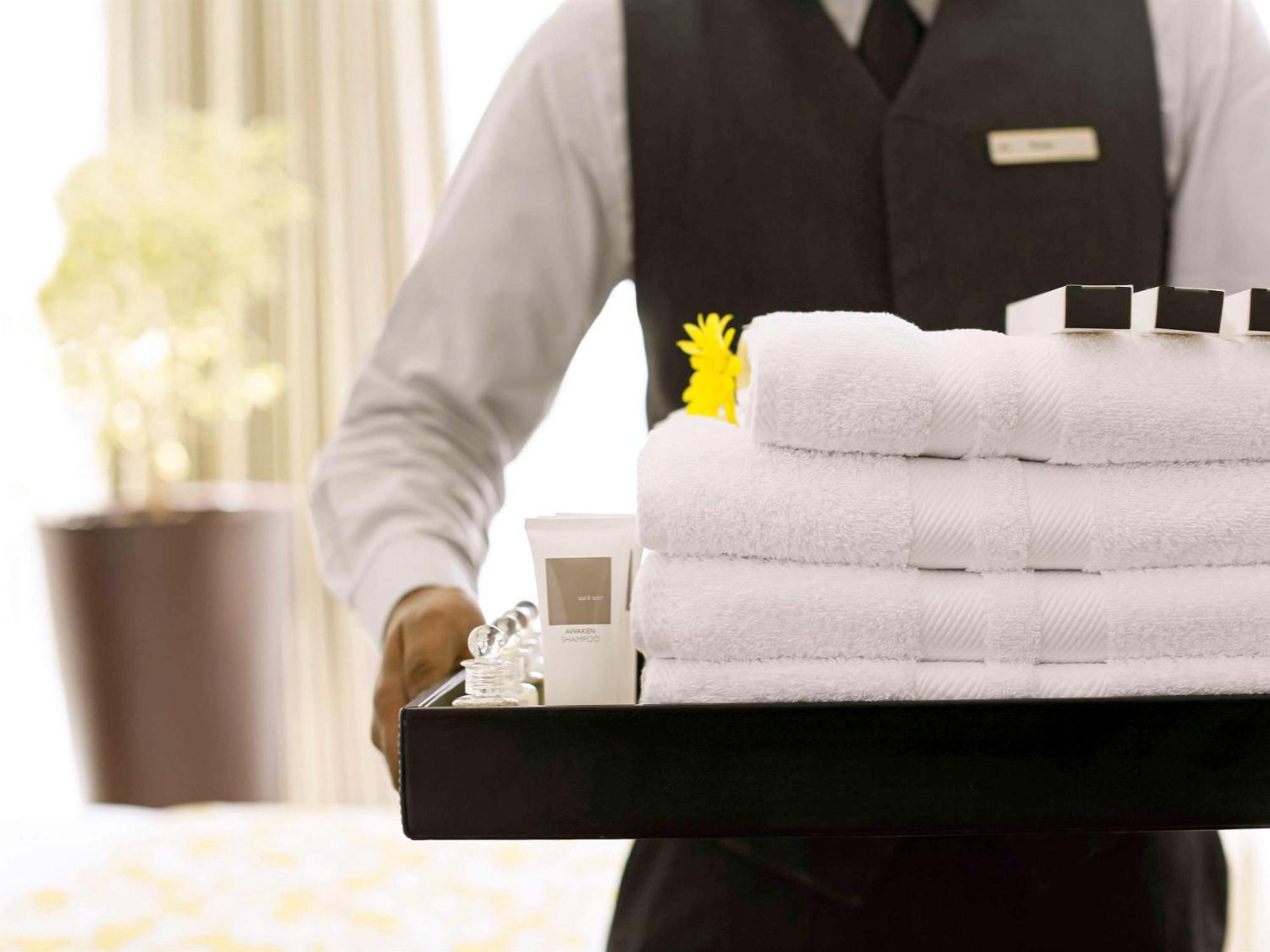 Swissotel Makkah La Meca Exterior foto The photo shows a person, likely a hotel staff member, holding a tray filled with neatly stacked white towels. The tray also contains a small tube of cream or lotion and several black and white square containers, possibly toiletries. A yellow flower 