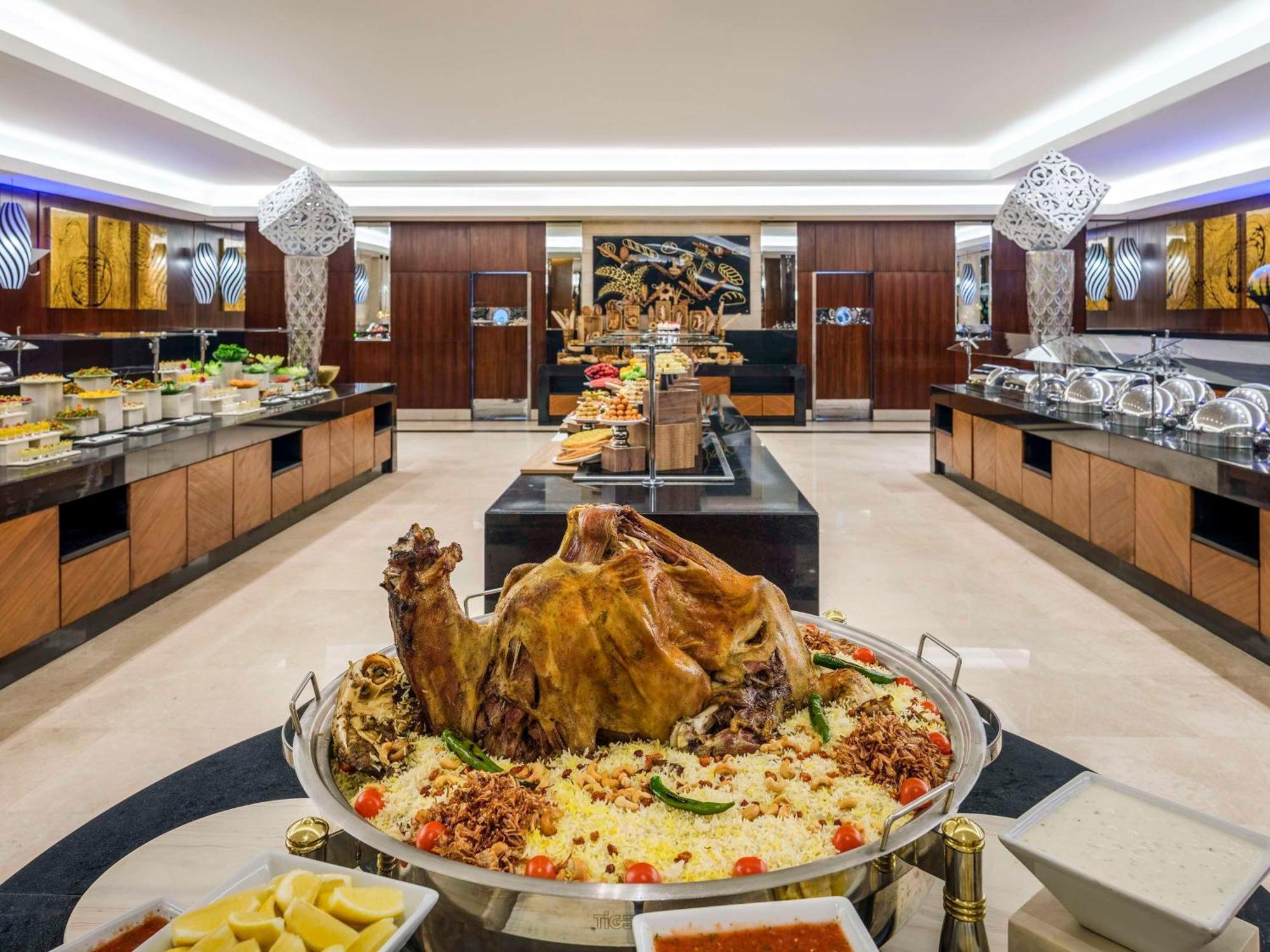 Swissotel Makkah La Meca Exterior foto The photo shows a lavish buffet setup in a restaurant or banquet hall. In the center, there is a large display of a roasted whole lamb or goat, garnished with herbs and served on a platter with rice and colorful accompaniments. Surrounding this cente