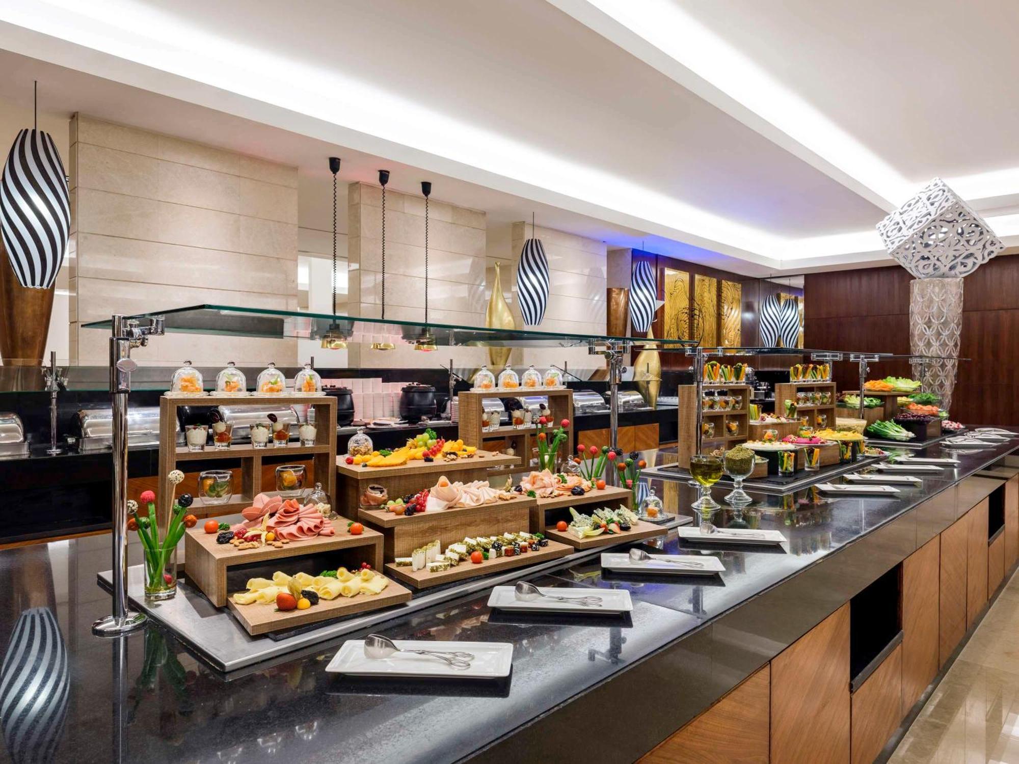 Swissotel Makkah La Meca Exterior foto The photo shows a buffet setup in a restaurant or hotel. There are several serving stations displaying a variety of foods. The setup includes tiered wooden racks filled with an assortment of dishes, likely featuring fruits, desserts, and appetizers. 