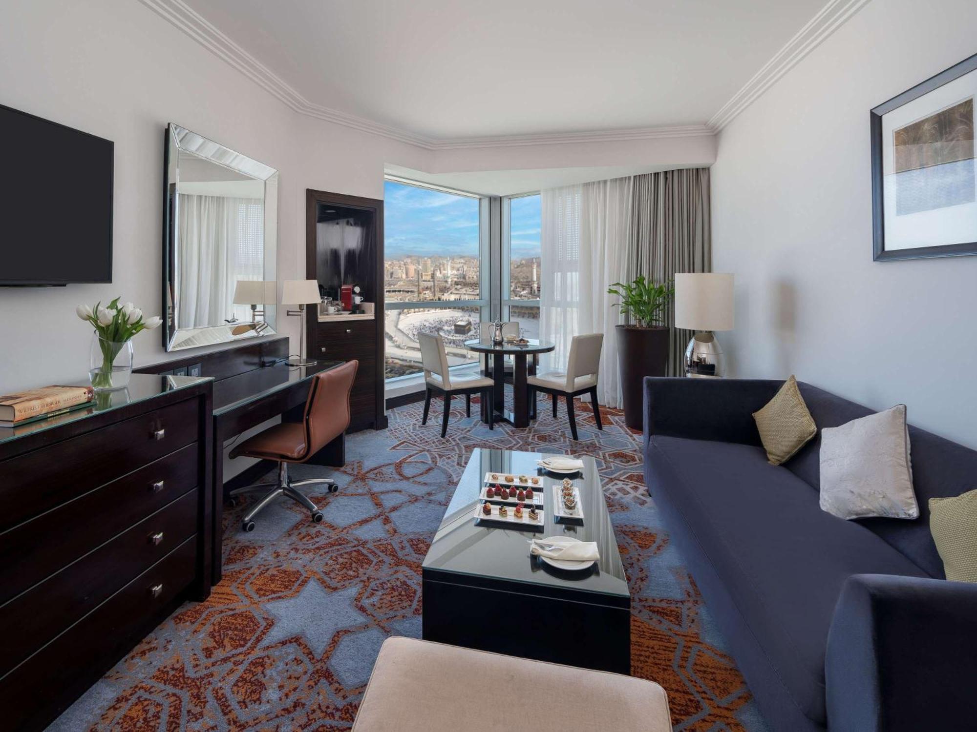 Swissotel Makkah La Meca Exterior foto The photo shows a hotel room featuring a modern and stylish design. There is a comfortable dark blue sofa and a coffee table in the center, along with light-colored accent pillows. Nearby, a small dining table with chairs is positioned near large win