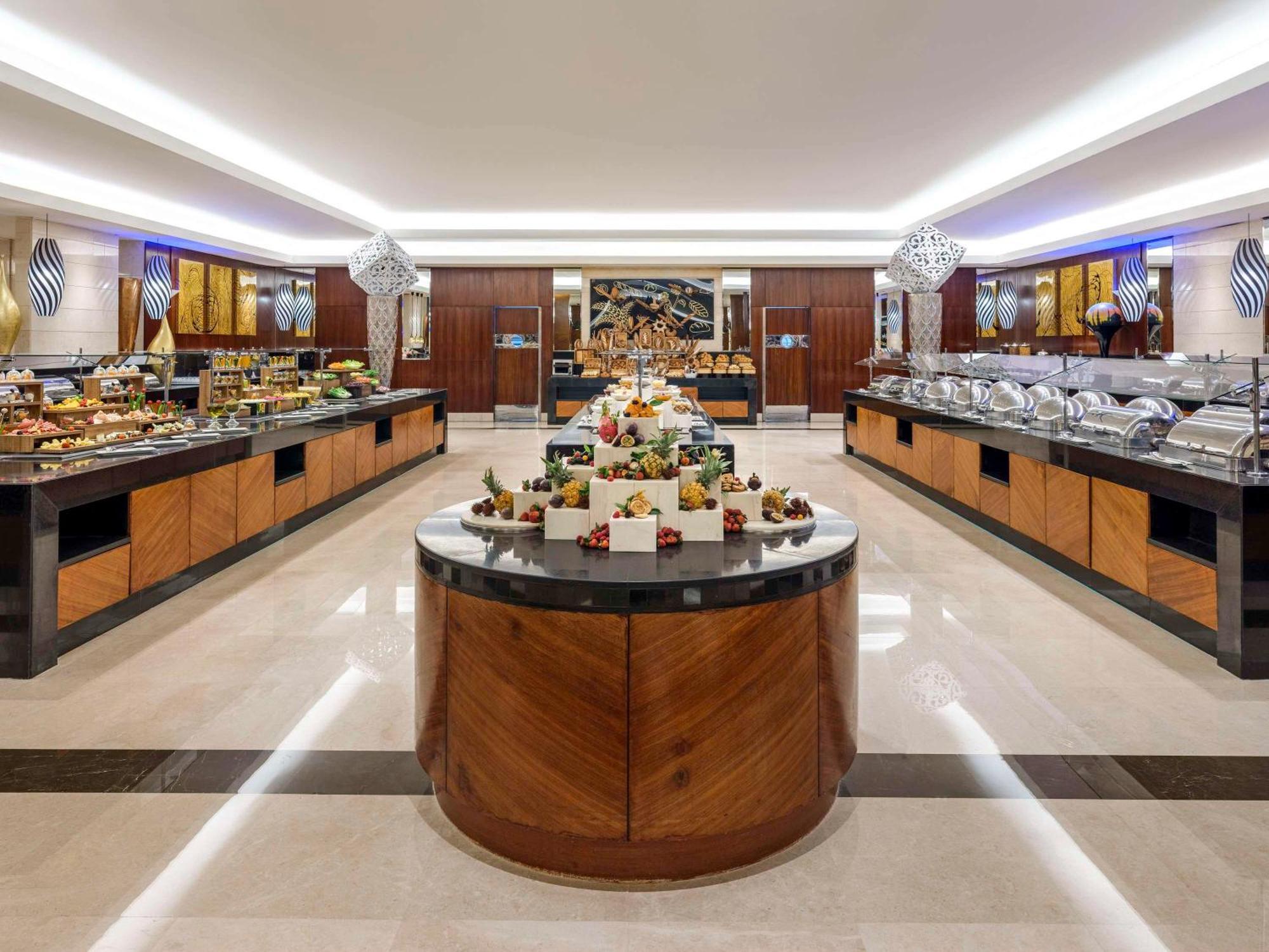 Swissotel Makkah La Meca Exterior foto The photo shows a spacious buffet area featuring elegantly arranged food displays. There are multiple food stations with wooden cabinetry, showcasing a variety of dishes. In the center, there is a round island display with an assortment of desserts o