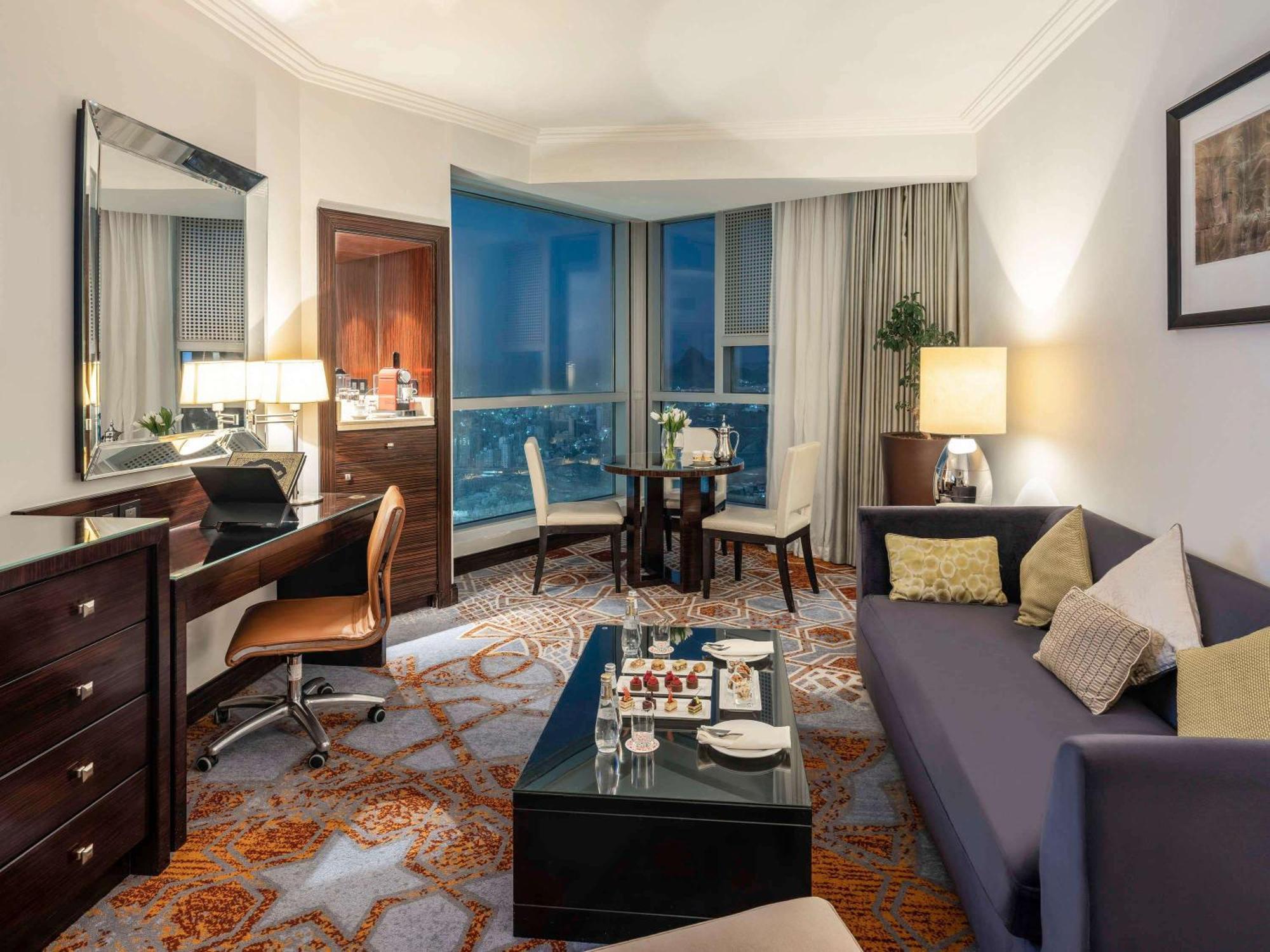 Swissotel Makkah La Meca Exterior foto The photo shows a well-furnished hotel suite or living area. It features a comfortable seating arrangement with a modern sofa and a coffee table. There is a dining area with a round table and chairs. On one side, there is a desk with a chair next to 