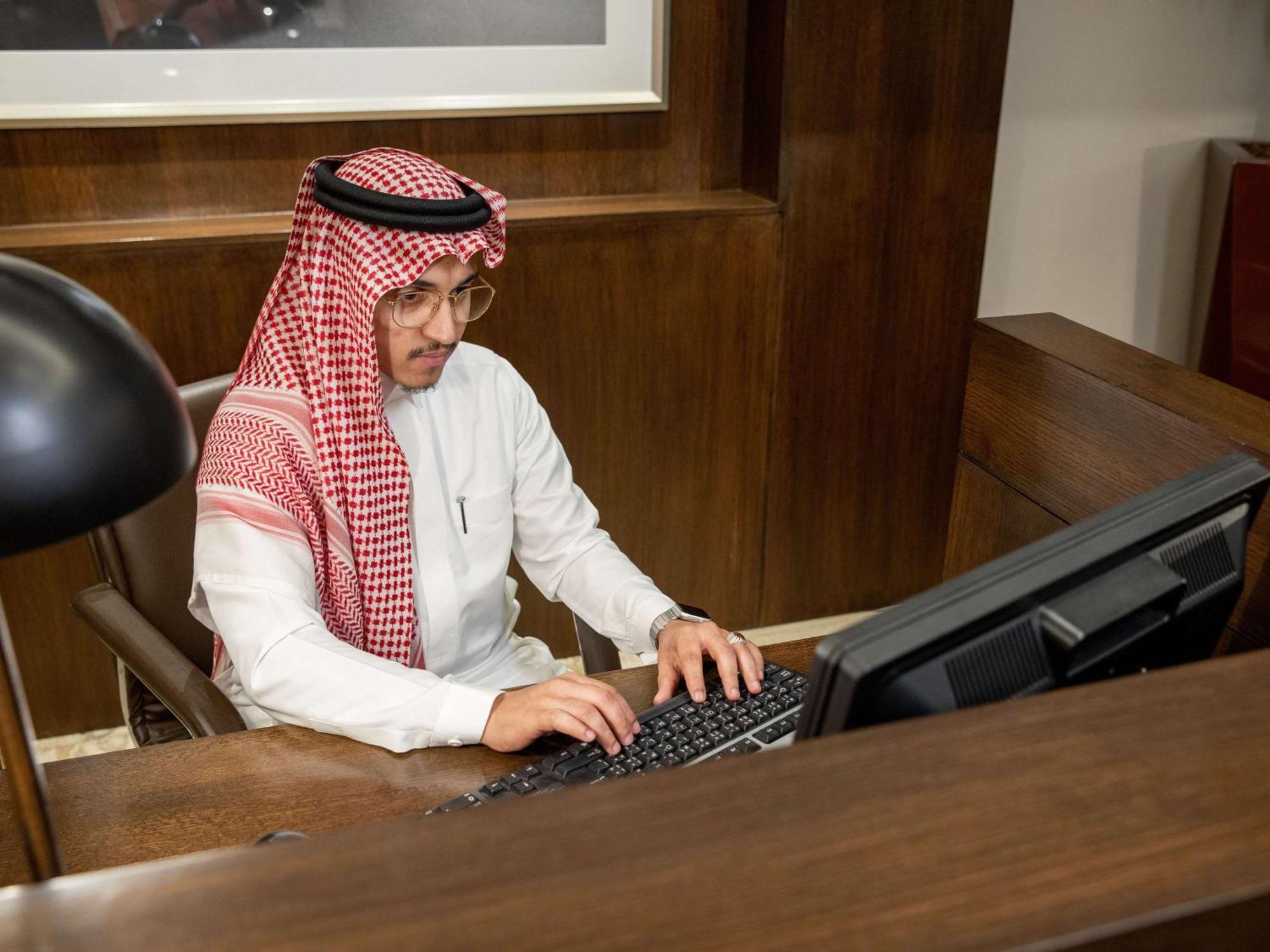 Swissotel Makkah La Meca Exterior foto The photo shows a man sitting at a desk, working on a computer. He is wearing traditional attire, including a white thobe and a red-and-white checked shemagh headdress. The setting appears to be an office, characterized by wooden furnishings and an o