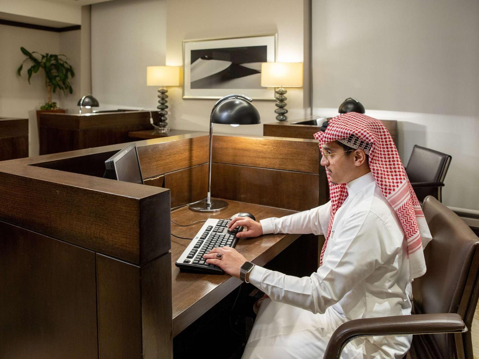 Swissotel Makkah La Meca Exterior foto The photo shows a man seated at a wooden desk in a modern office setting. He is dressed in traditional attire, which includes a white thobe and a red and white checked headscarf. The man is focused on typing on a keyboard, and there is a desk lamp pr