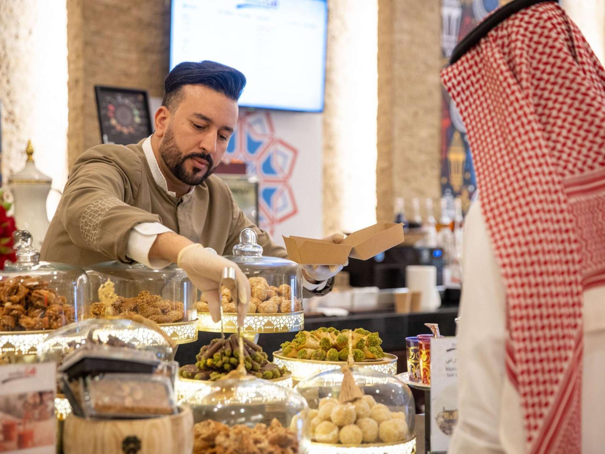 Swissotel Makkah La Meca Exterior foto The photo shows a scene in a food establishment, likely a market or café. A man dressed in a traditional outfit is serving food from a display of various dishes. He is wearing gloves and is placing items into a box for a customer. The display feature