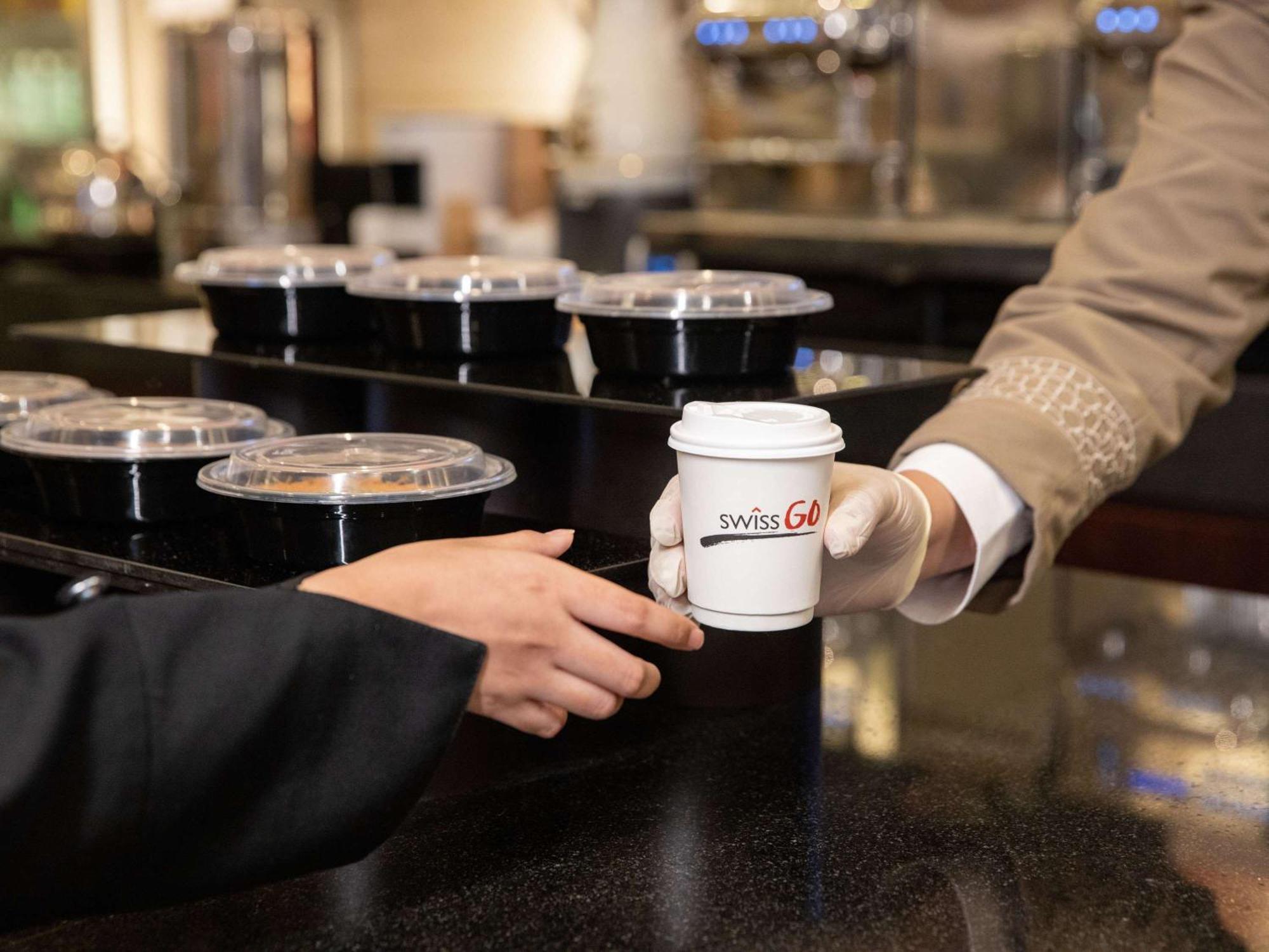 Swissotel Makkah La Meca Exterior foto The image shows a scene at a counter where a person is handing over a cup of coffee with a logo that reads "SwissGo." The person receiving the coffee is wearing a black jacket, while the one serving it is dressed in a beige uniform. In the background