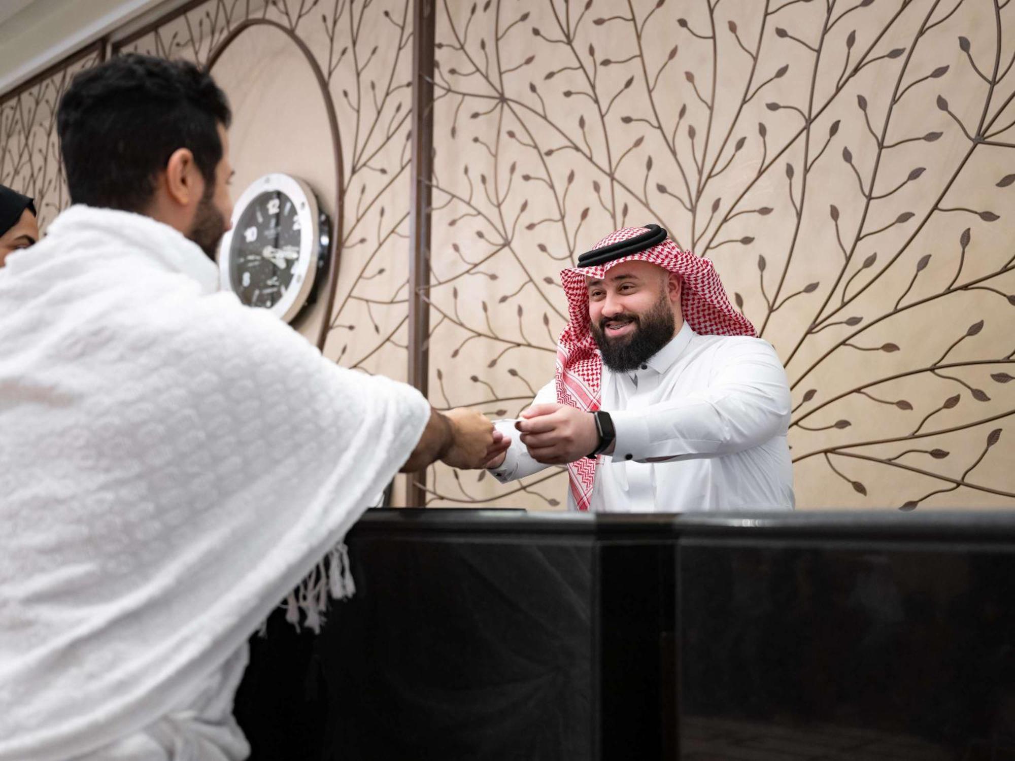 Swissotel Makkah La Meca Exterior foto The image shows two individuals engaged in a handshake. One person, dressed in white traditional attire, appears to be greeting the other, who is wearing a thobe and a traditional headscarf (ghutrah). The background features decorative wall art and a